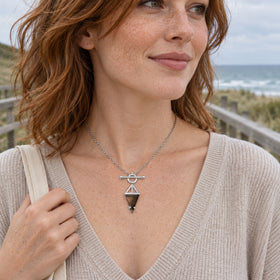 A freckled woman with auburn hair wears the silver triangular smoky quartz necklace with a pale knit top beside a coastal boardwalk.