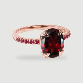 An oval faceted red gemstone sits in a rose gold vermeil ring with red stone shoulders.