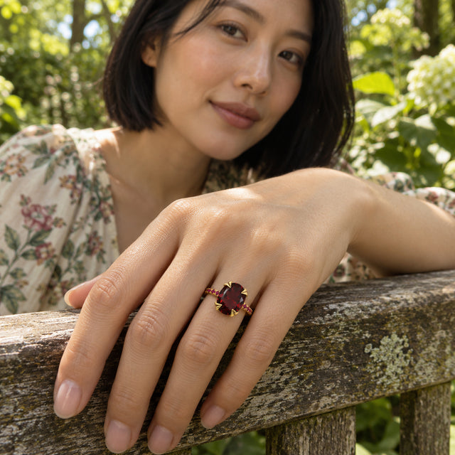 A woman wears a yellow gold vermeil ring with an oval red stone while resting her hand on a weathered garden bench.