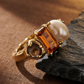 The gold ring rests against dark slate on a textured tabletop, showing the raised amber stone and white pearl.