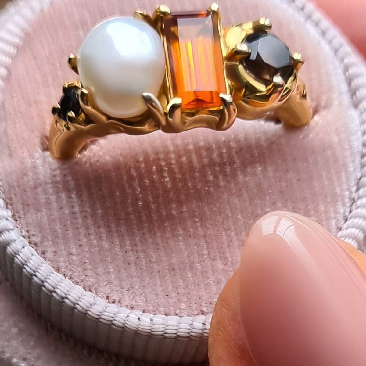 The gold ring is displayed on pale pink velvet with its amber rectangular stone, white pearl, and round dark accents visible.