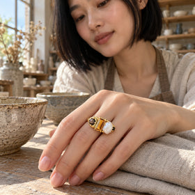 The gold ring is worn on a relaxed hand beside ceramic bowls and folded linen at a sunlit workbench.