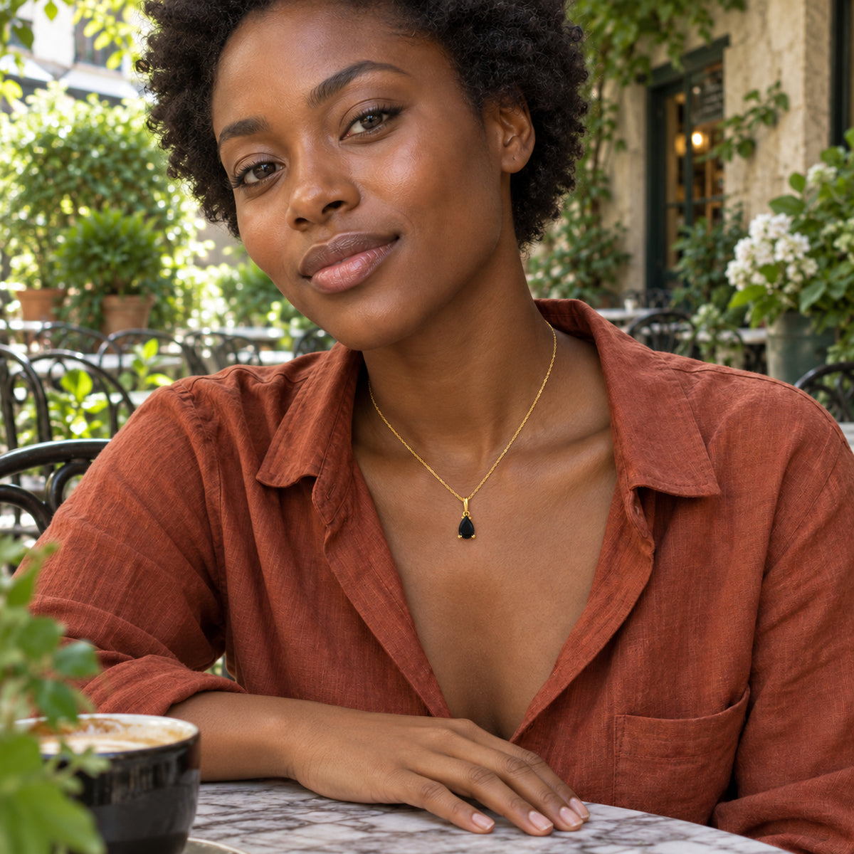 A woman in a rust-colored shirt wears a small black pear-shaped pendant on a yellow gold-colored chain at an outdoor cafe.