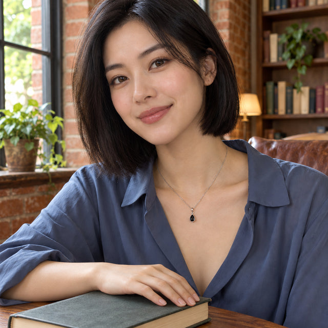A woman in a slate blue blouse wears a black pear-shaped pendant on a silver-colored chain beside a window and bookshelves.