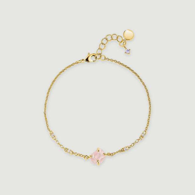 A yellow gold tone chain bracelet forms a loop with a raw pink stone and small clear accents on a pale grey surface.