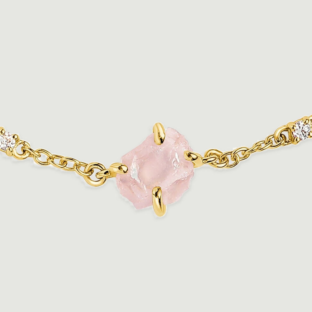 A close view shows the rough pink crystal held by yellow gold tone prongs beside two clear stones and oval chain links.