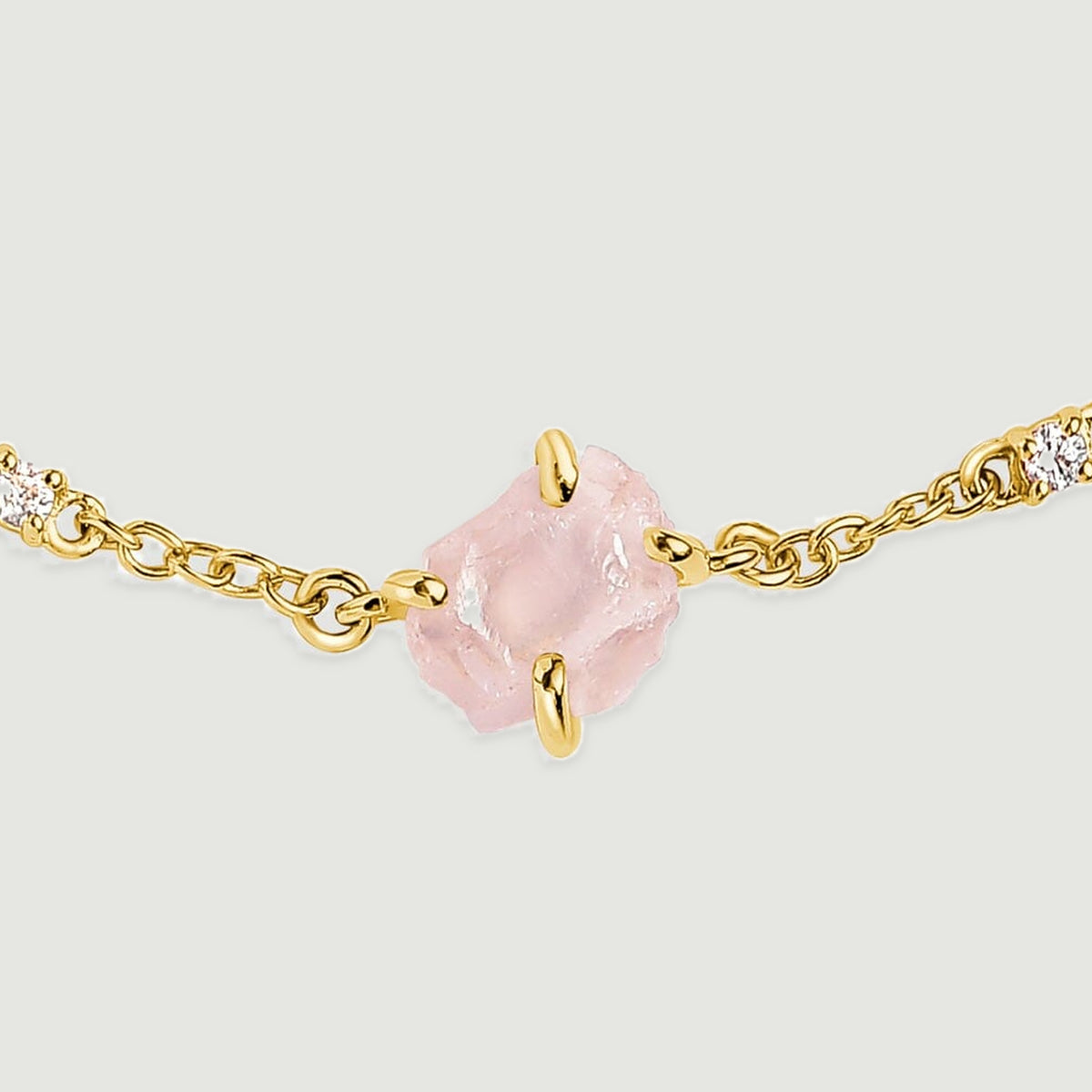 A close view shows the rough pink crystal held by yellow gold tone prongs beside two clear stones and oval chain links.