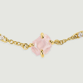 A close view shows the rough pink crystal held by yellow gold tone prongs beside two clear stones and oval chain links.