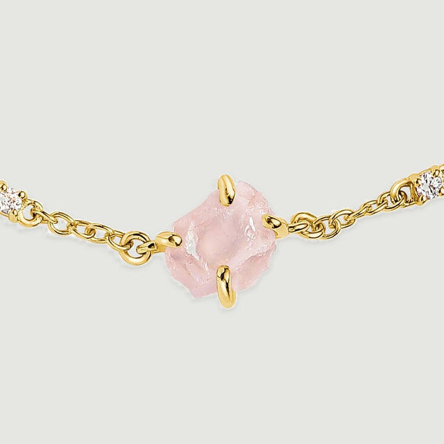 A close view shows the rough pink crystal held by yellow gold tone prongs beside two clear stones and oval chain links.