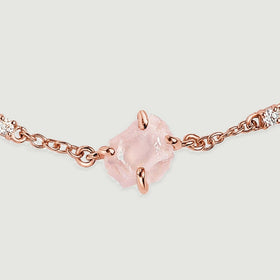 The uneven surface of a pale pink stone is shown between polished rose gold tone prongs and clear round accents.