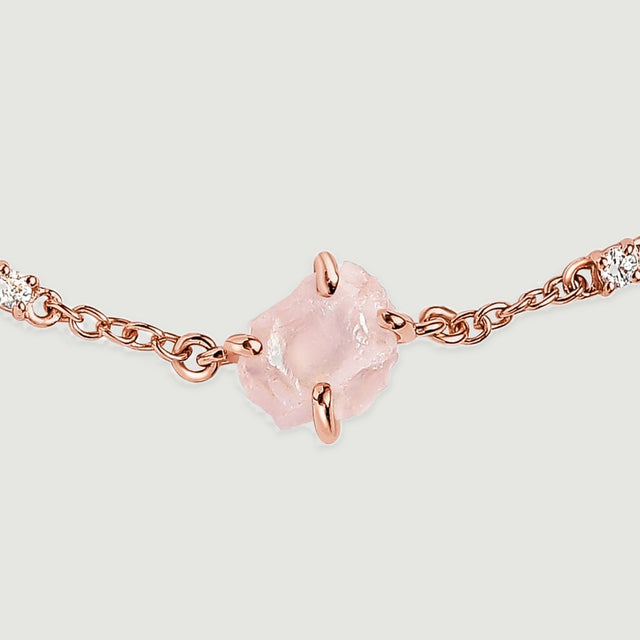 The uneven surface of a pale pink stone is shown between polished rose gold tone prongs and clear round accents.