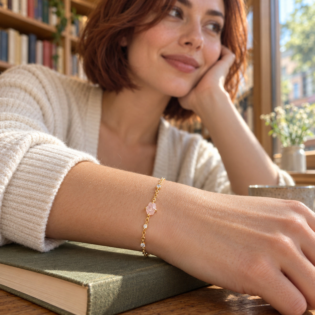 A yellow gold tone bracelet with a raw pink stone is worn on a wrist resting on a closed book beside a sunny window.
