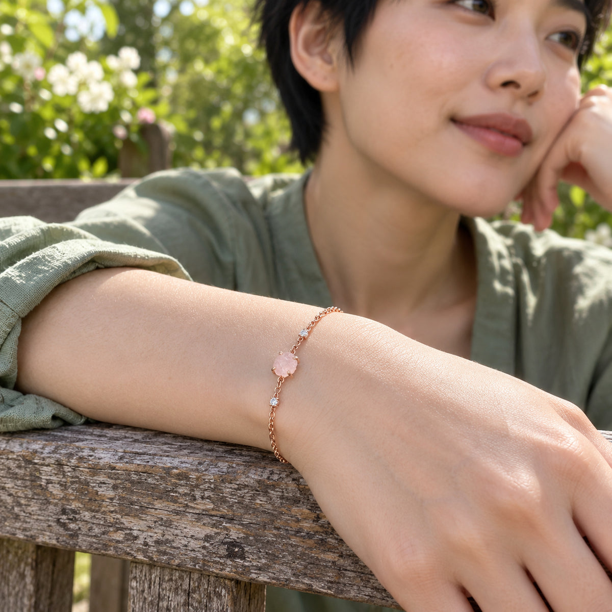 A rose gold tone chain and pale pink crystal circle a wrist resting on a weathered wooden bench in a leafy garden.