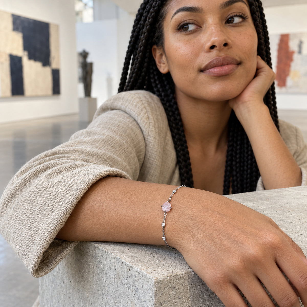 A silver tone bracelet with a pink crystal and clear accents is worn above a stone ledge with framed artwork behind the wearer.
