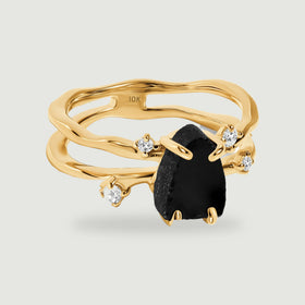 A yellow gold ring with a rough black stone and four clear accents rests on a pale grey background.