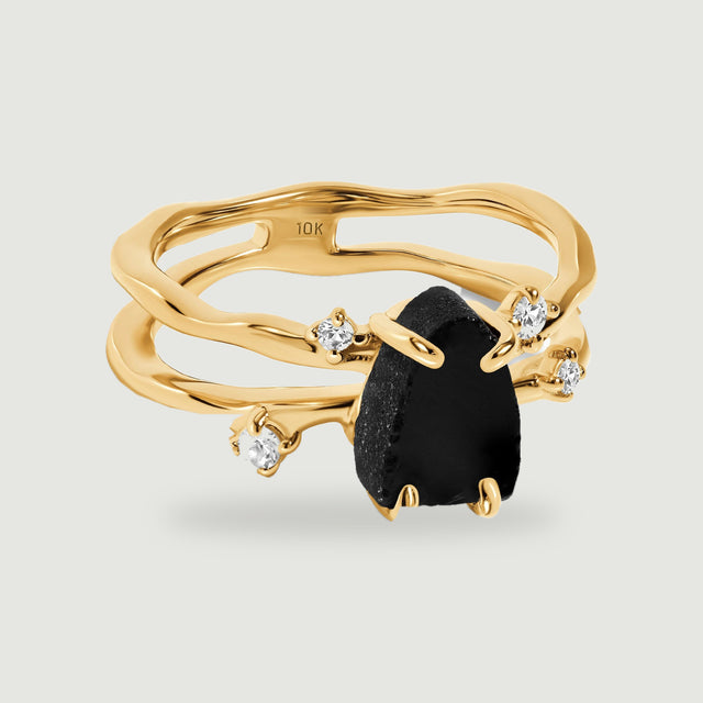 A yellow gold ring with a rough black stone and four clear accents rests on a pale grey background.