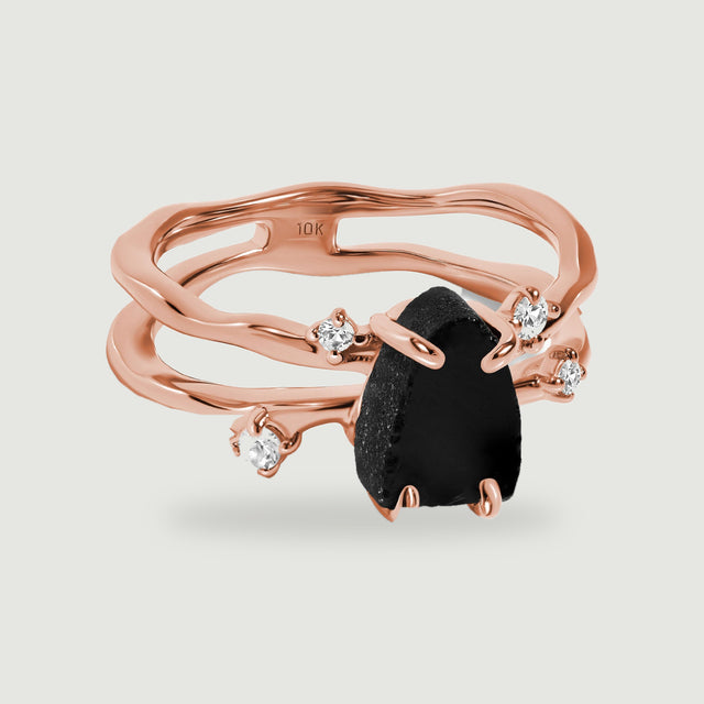 A rose gold split band surrounds a dark irregular stone with four small clear stones on pale grey.