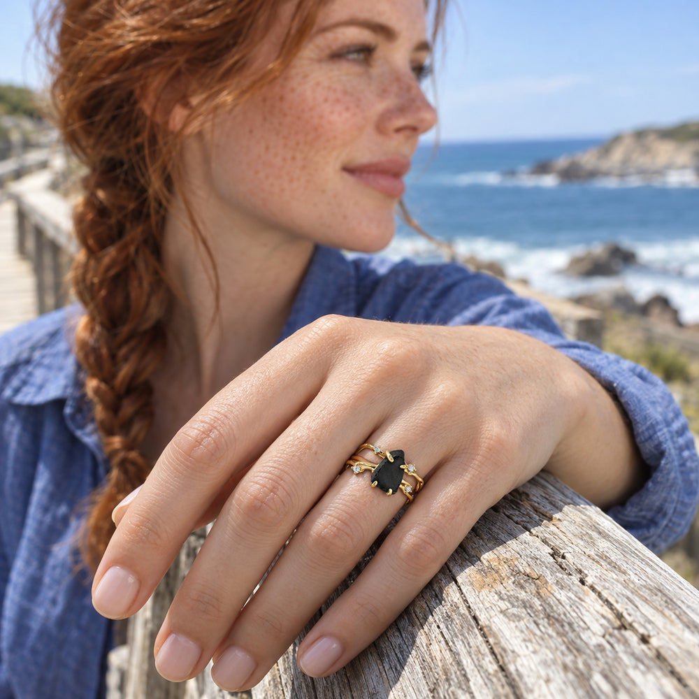 A yellow gold ring with a black stone is worn on a hand resting on a wooden seaside railing.