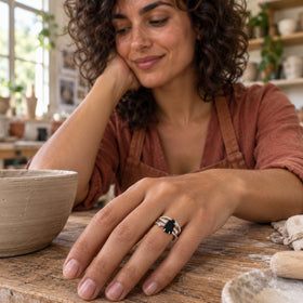 A white gold ring with a dark irregular stone is worn on a hand resting on a timber bench beside a clay bowl.