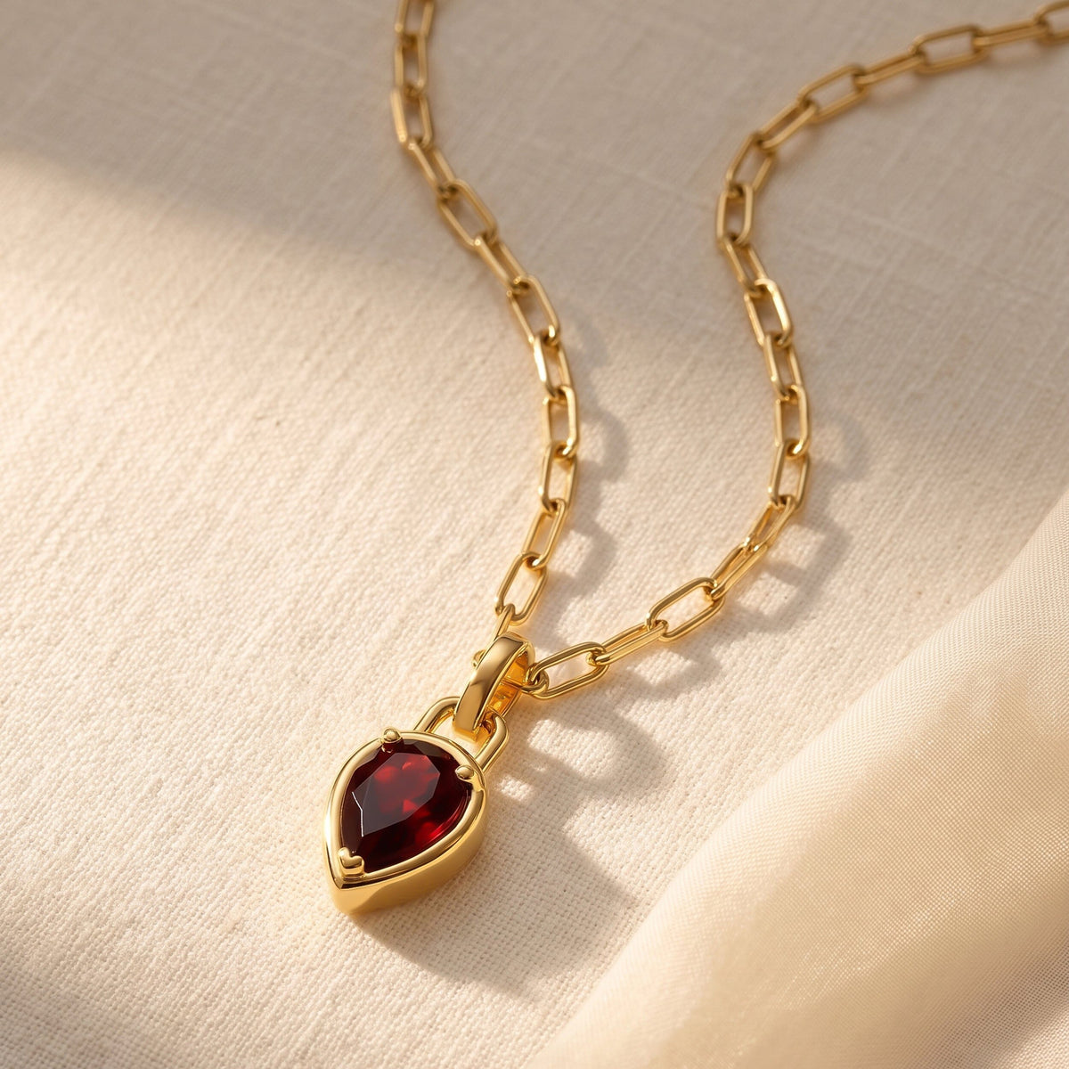 A yellow gold-tone chain and red garnet pendant lie diagonally across softly folded cream linen.