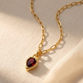 A yellow gold-tone chain and red garnet pendant lie diagonally across softly folded cream linen.