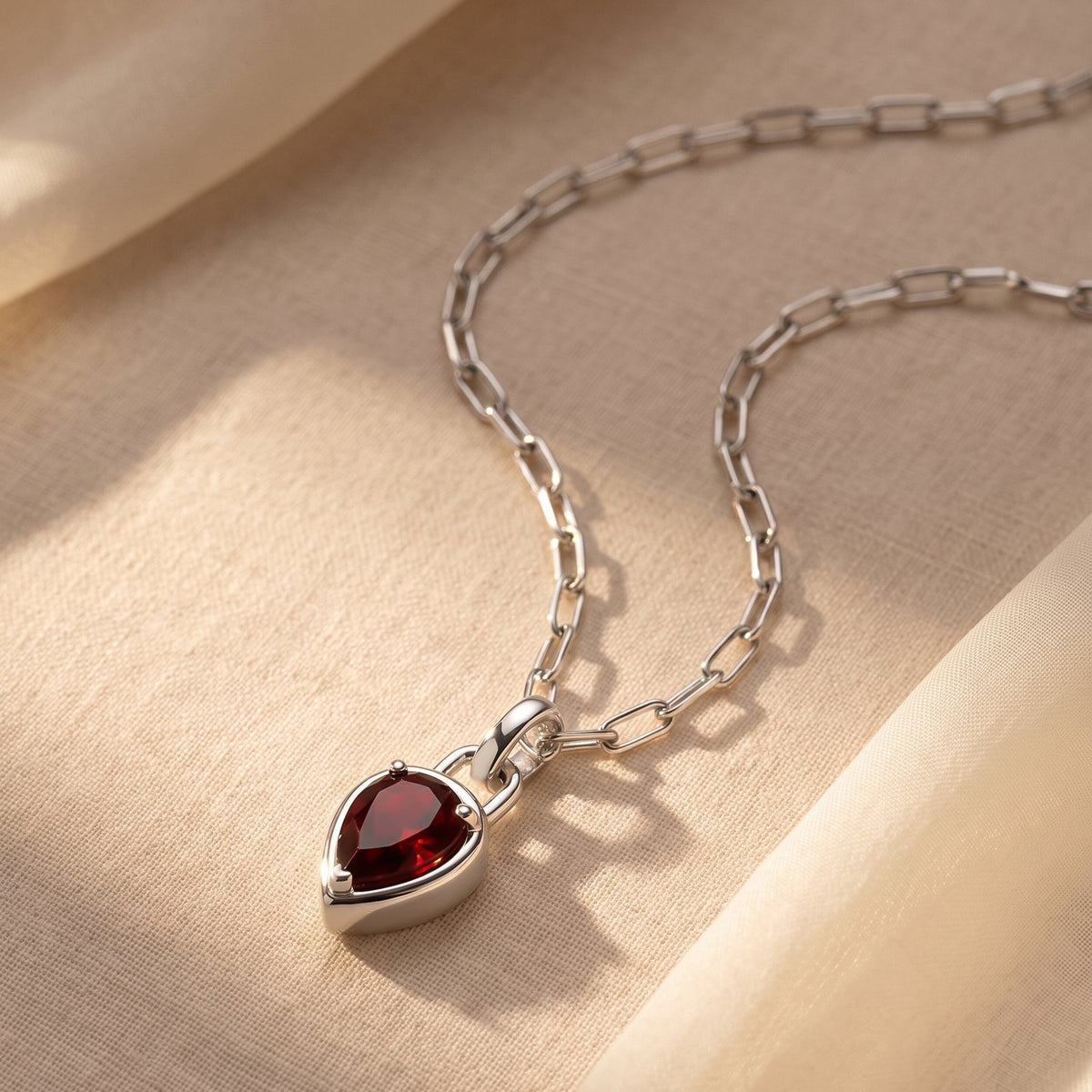 A silver-tone link necklace rests on cream fabric with the red pendant near the lower edge.