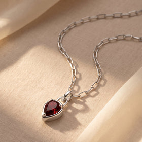 A silver-tone link necklace rests on cream fabric with the red pendant near the lower edge.