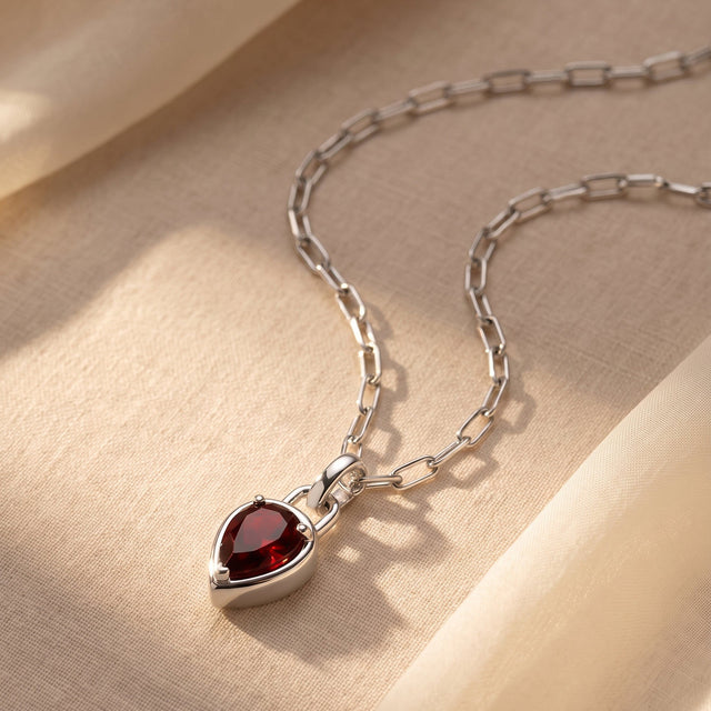 A silver-tone link necklace rests on cream fabric with the red pendant near the lower edge.