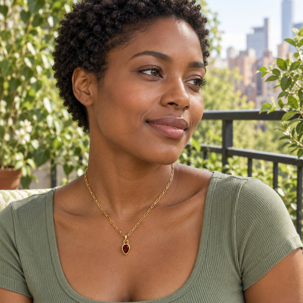 A woman with short curls wears a gold-tone red garnet necklace and a sage green top on a leafy rooftop terrace.