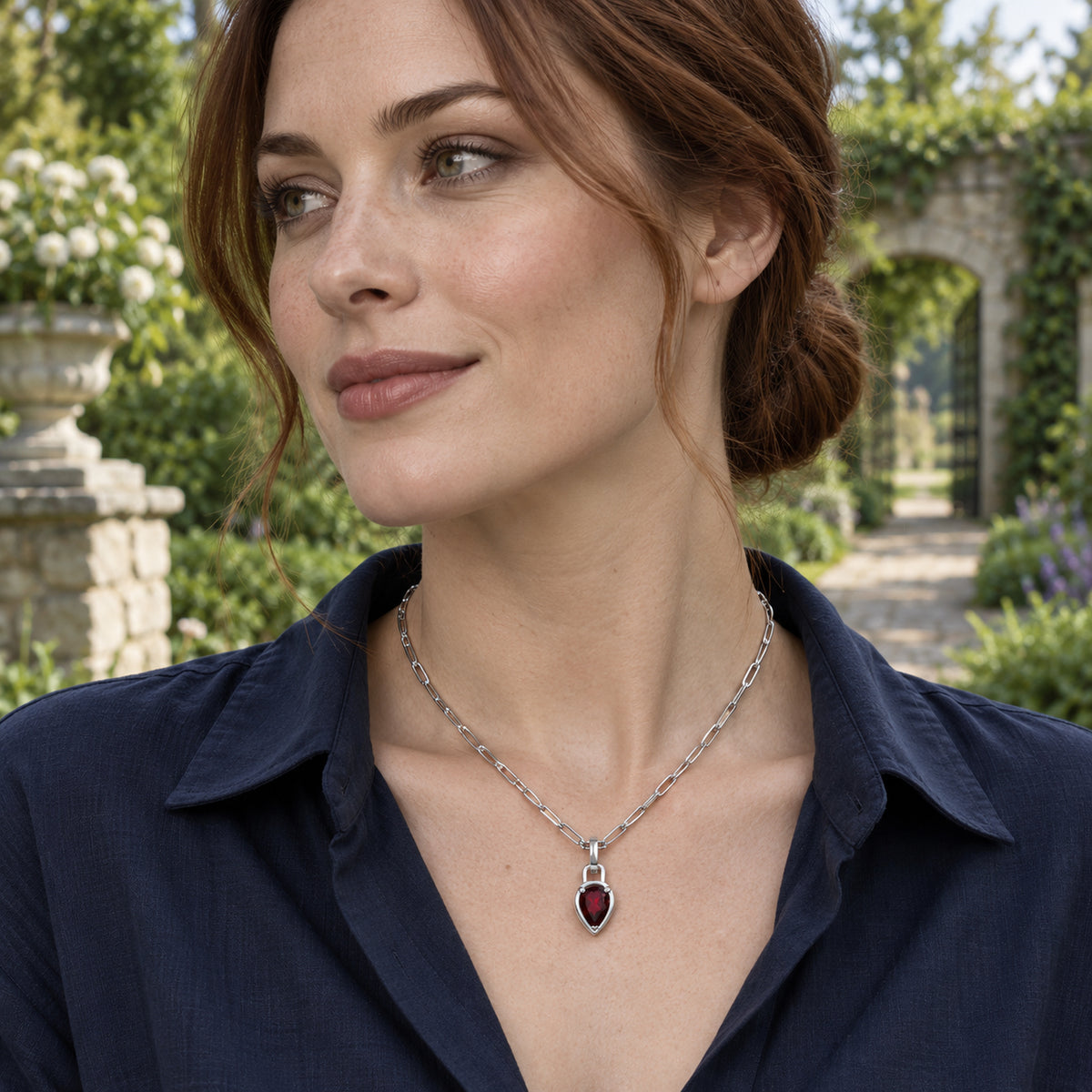 An auburn-haired woman wears a silver-tone garnet pendant necklace with a navy shirt in a sunlit garden.