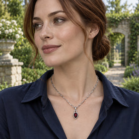 An auburn-haired woman wears a silver-tone garnet pendant necklace with a navy shirt in a sunlit garden.
