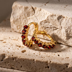 Two yellow gold plated garnet hoops rest beside a pale textured stone block.