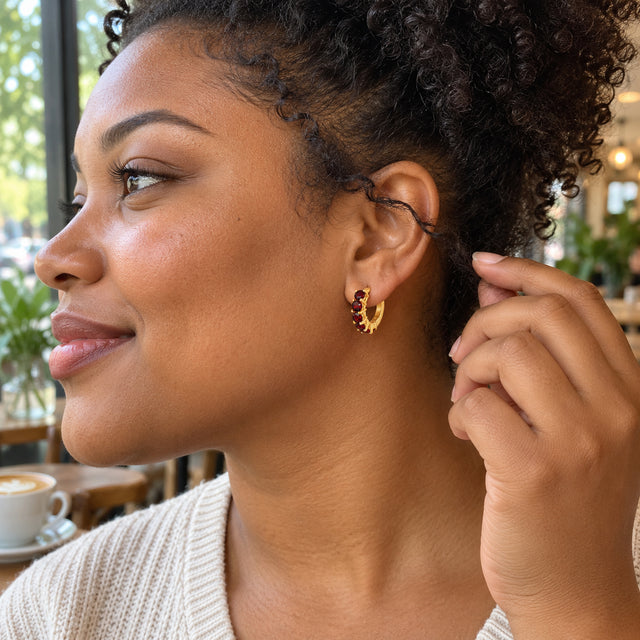 A woman with curly hair wears a yellow gold plated garnet hoop beside a cafe window.