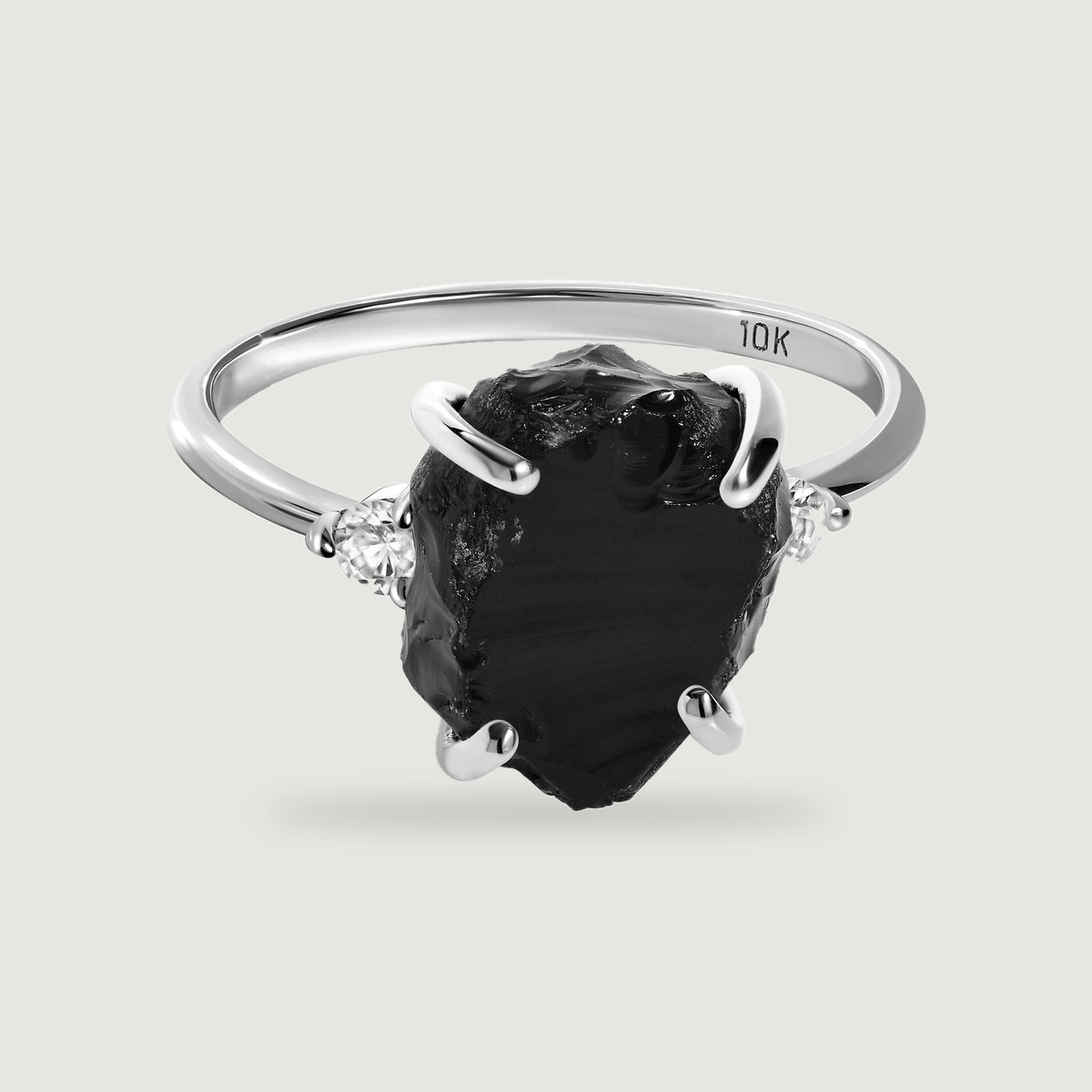 A white gold ring with a raw black stone and two clear side stones rests on a pale gray surface.