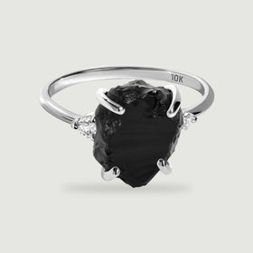 A white gold ring with a raw black stone and two clear side stones rests on a pale gray surface.