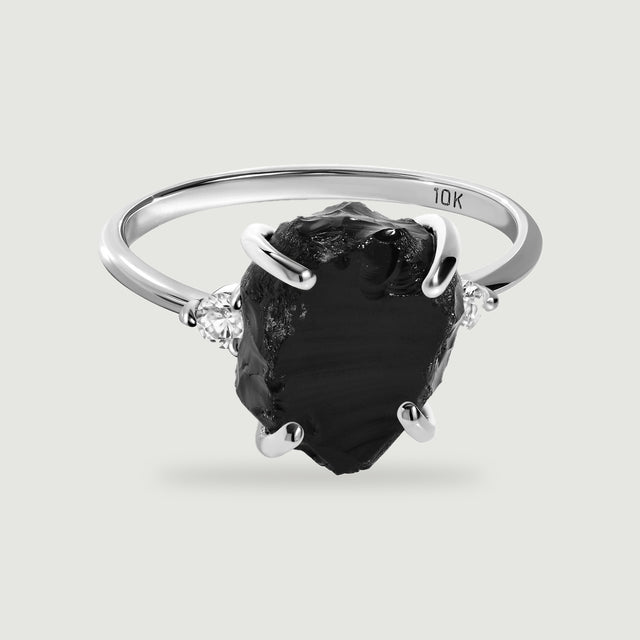 A white gold ring with a raw black stone and two clear side stones rests on a pale gray surface.