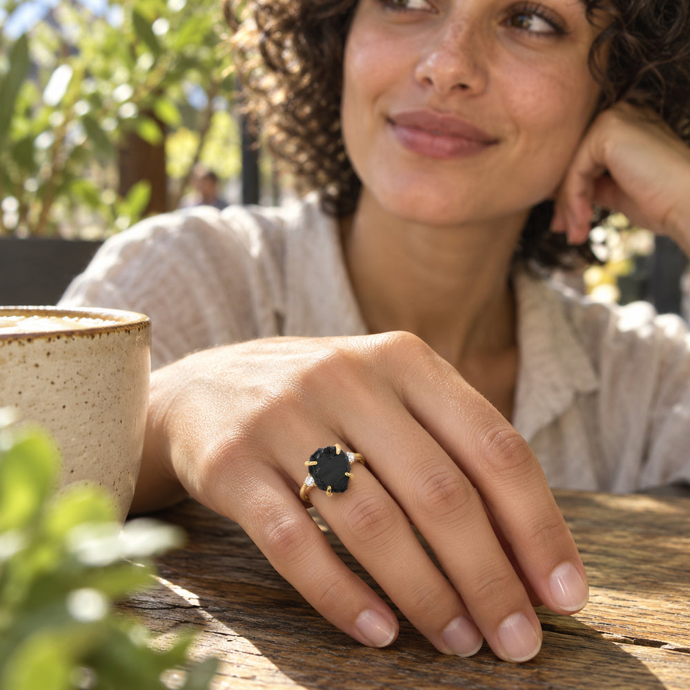 A yellow gold ring with a rough black center stone is worn on a hand resting beside a ceramic cup at a wooden cafe table.