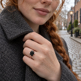 A rose gold ring with a black center stone and two clear side stones is worn on a freckled hand holding a dark wool coat.