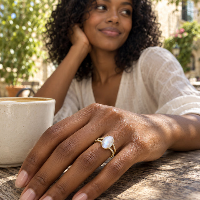 A yellow gold moonstone ring is worn on a hand beside a ceramic cup at a sunlit courtyard table.