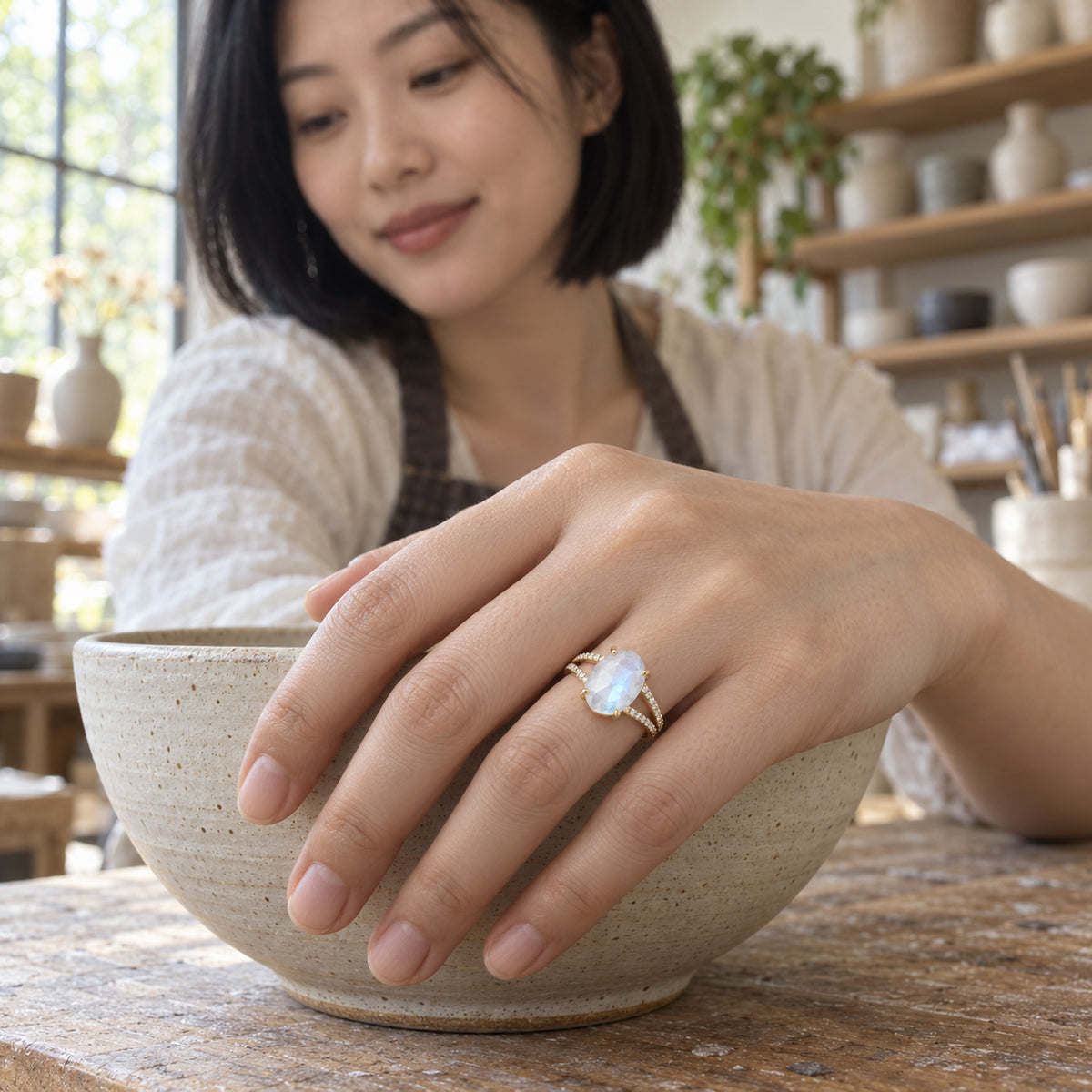 The oval moonstone ring is shown on a hand resting over a ceramic bowl in a pottery studio.