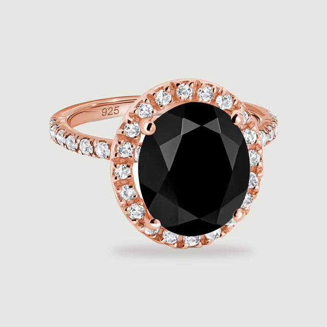 An oval faceted black ring with a clear stone halo and rose gold band rests on a light gray background.