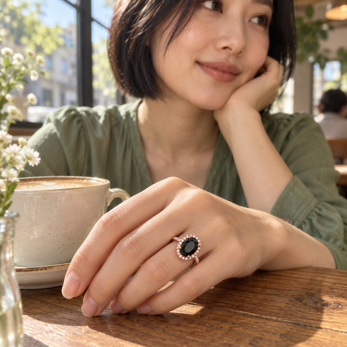A woman in a green blouse wears the black oval rose gold ring on her hand beside a coffee cup on a wooden cafe table.