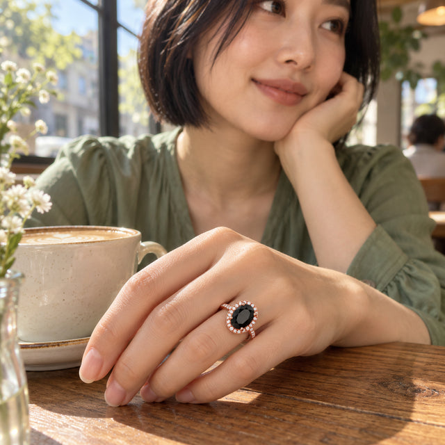 A woman in a green blouse wears the black oval rose gold ring on her hand beside a coffee cup on a wooden cafe table.
