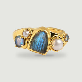 A yellow gold vermeil ring with a raw blue labradorite center and pearl accents casts a soft shadow on a gray background.