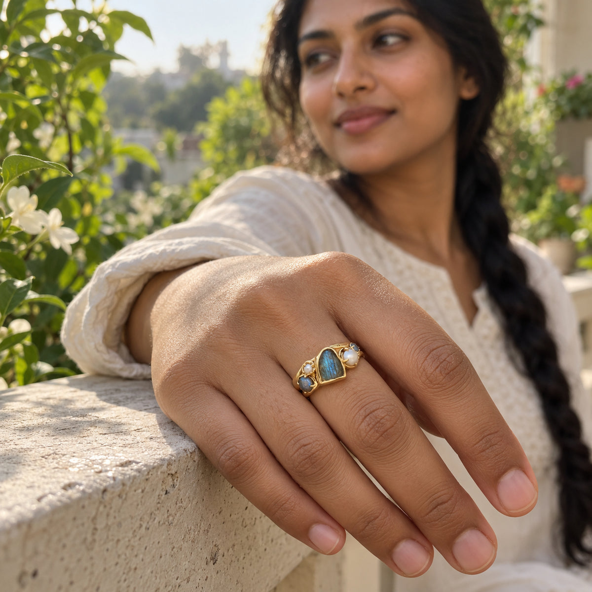A woman wears the yellow gold vermeil labradorite and pearl ring while resting her hand on a stone railing in a leafy garden.
