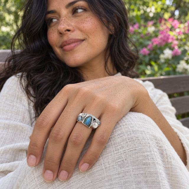 A woman wears the silver labradorite and pearl ring on her hand over an ivory linen sleeve beside a flowering garden and wooden bench.
