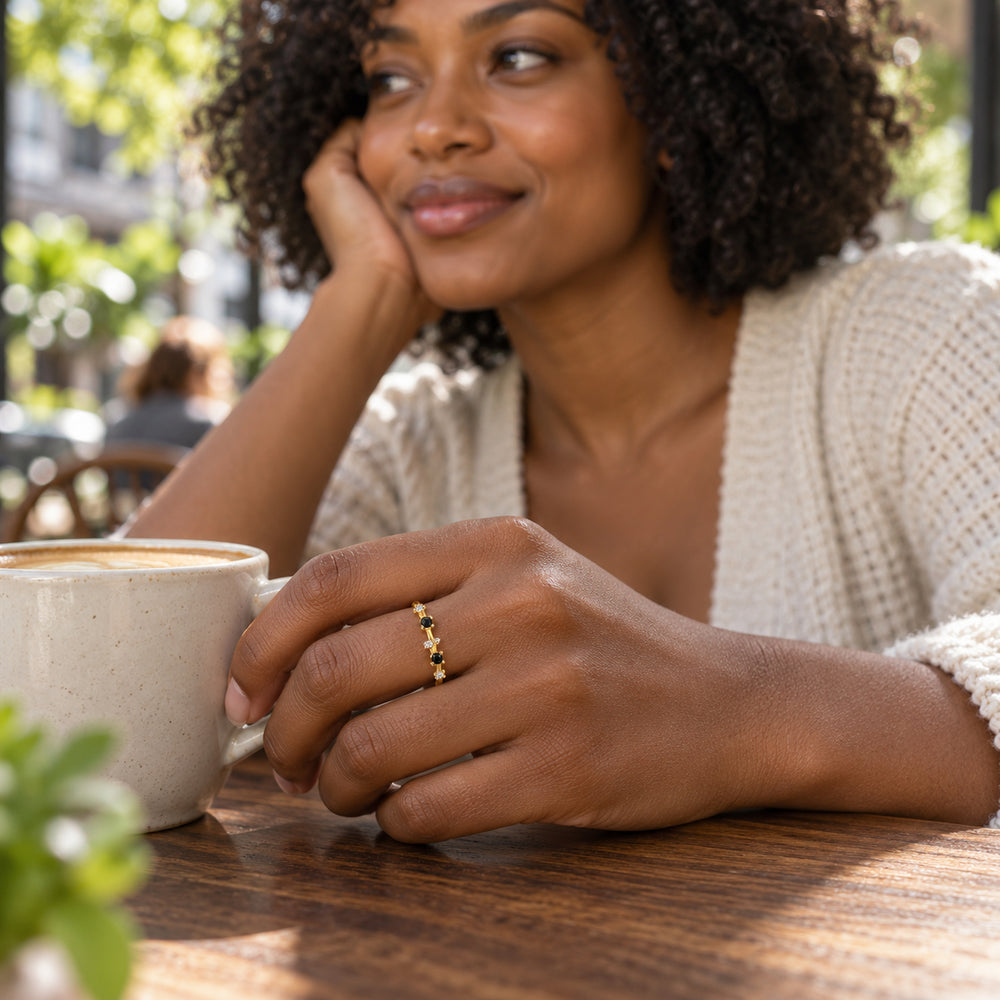A woman with curly hair wears a yellow gold vermeil ring with black and clear stones beside a coffee cup at an outdoor cafe.