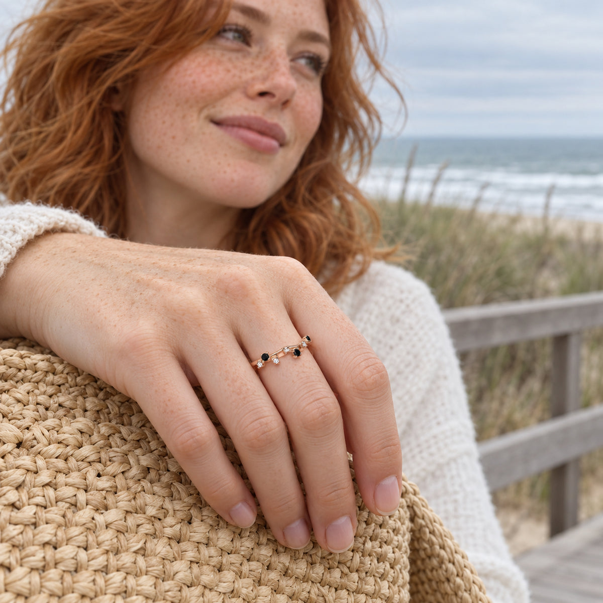 A freckled red-haired woman shows a rose gold vermeil ring with black and clear stones while resting her hand on a woven bag beside the sea.