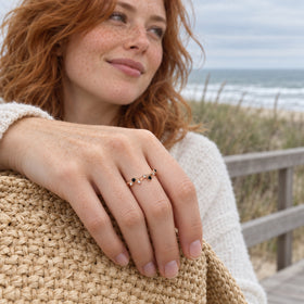 A freckled red-haired woman shows a rose gold vermeil ring with black and clear stones while resting her hand on a woven bag beside the sea.