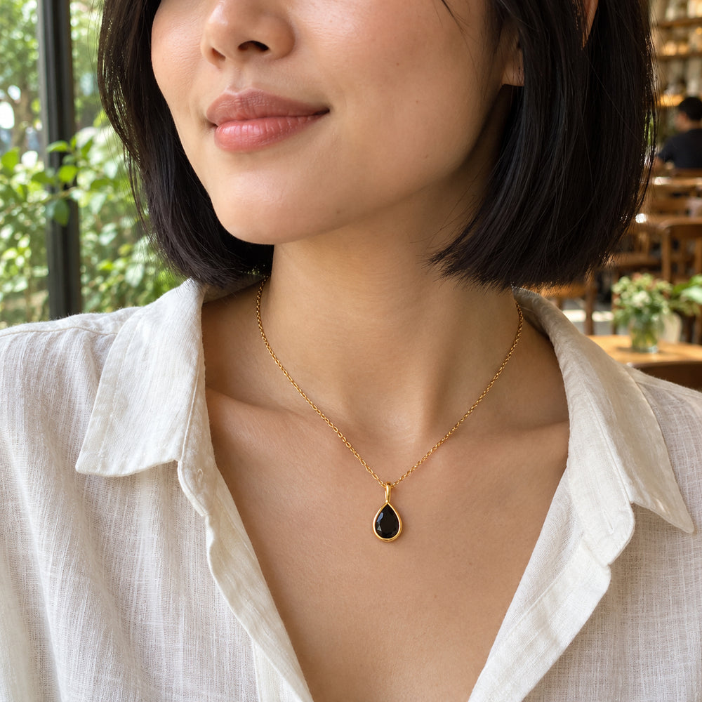 A woman with a black bob wears a yellow gold necklace with a black pear-shaped pendant beside a leafy cafe window.